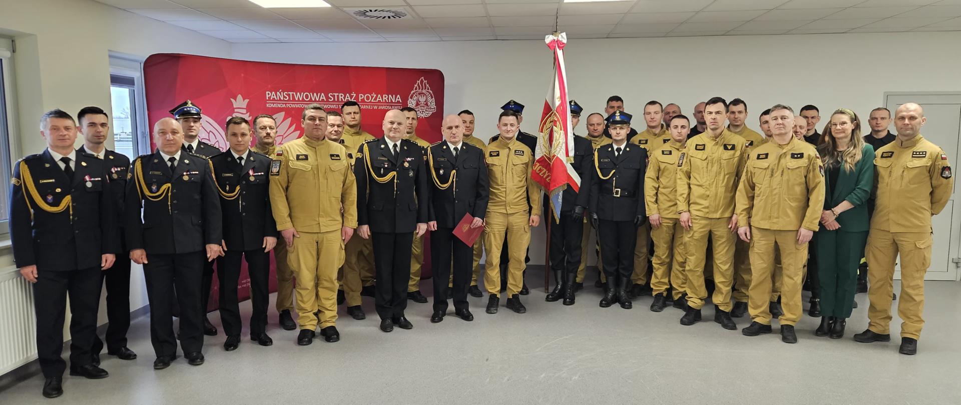 Grupa funkcjonariuszy Państwowej Straży Pożarnej w umundurowaniu służbowym i galowym, ustawionych w kilku rzędach do wspólnego zdjęcia, na tle ścianki z napisem „Państwowa Straż Pożarna