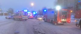 Na zdjęciu widać samochody pożarnicze oraz karetki pogotowia, które obsługują zdarzenie na drodze. Na jezdni leży śnieg zaczyna robić się zmierzch. Droga jest całkowicie zablokowana na jezdni widać porozrzucane plastikowe elementy z rozbitych aut. W tle po prawej stronie widać wieżowce