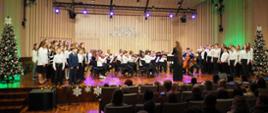 Chór dziecięcy oraz orkiestra dziecięca smyczkowa z dyrygentem na scenie w sali koncertowej, po bokach dekoracyjne świąteczne choinki, w tle napis Koncert Noworoczny, przed sceną publiczność.