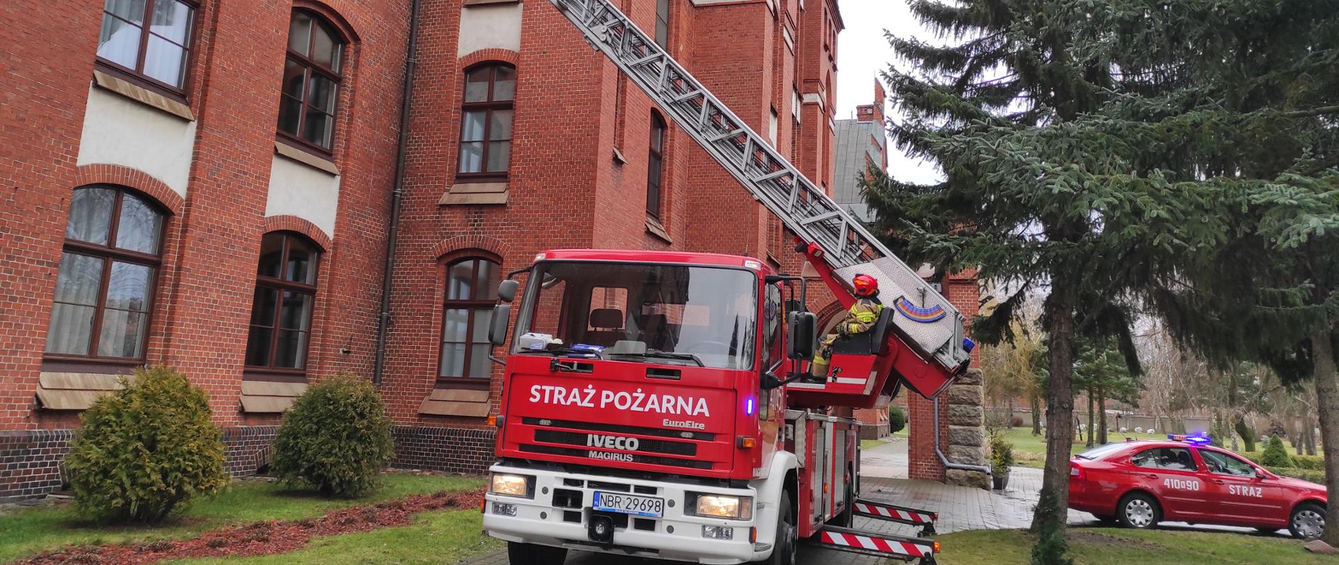 Na tle dużego czerwonego budynku stoi rozstawiana auto drobina strażacka, w koszu strażak.