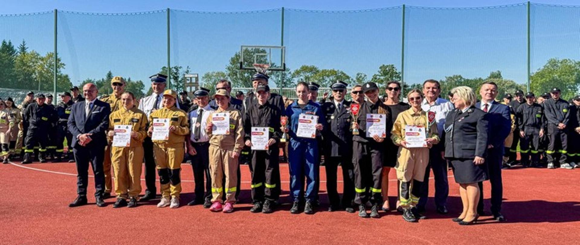 Na zdjęciu widać grupę osób ustawionych w rzędzie na czerwonym boisku sportowym. Pośrodku znajdują się osoby, które otrzymały nagrody lub dyplomy — część z nich jest ubrana w beżowe lub granatowe stroje strażackie, a część w stroje galowe lub eleganckie ubrania. Wszyscy trzymają w rękach dyplomy. W tle, po obu stronach centralnej grupy, widać duży szpaler strażaków w czarnych uniformach. Za nimi znajduje się zielona siatka sportowa, a w oddali widać drzewa i błękitne niebo.