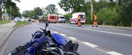 Na poboczu leży granatowy motocykl marki Yamaha. W oddali znajdują się 2 karetki pogotowia, wóz Straży Pożarnej oraz samochód Policji. Obok pojazdów znajduje się Zespół Ratownictwa Medycznego oraz strażacy.