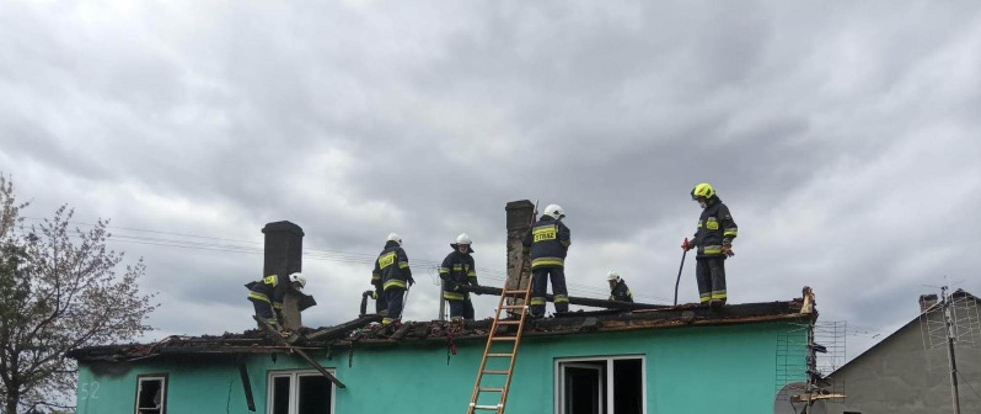Zdjęcie przedstawia strażaków pracujących na dachu spalonego budynku mieszkalnego. Na pierwszym planie widać strażaka przytrzymującego drabinę nasadkową przystawioną do ściany budynku.