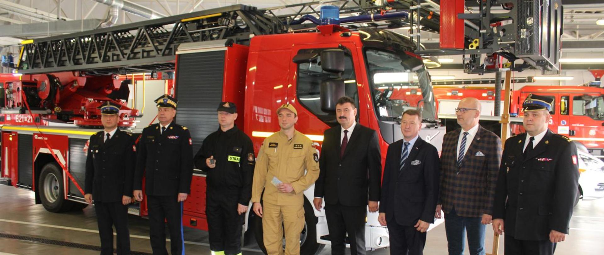 Zdjęcie przedstawia Posła, Komendantów PSP oraz zaproszonych gości przekazujących nowy pojazd. W tle samochód specjalny z drabiną mechaniczną, w remizie.