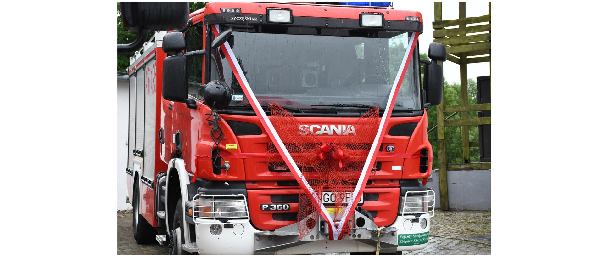 Zdjęcie przedstawia udekorowany ciężki samochód ratowniczo-gaśniczy podczas uroczystości jego przekazania do jednej z jednostek Ochotniczych Straży Pożarnych