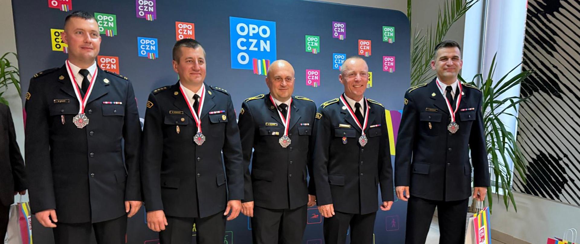 5 strażaków w umundurowaniu bez nakrycia głowy pozuje do zdjęcia z medalami. W tle ścianka z napisami Opoczno. Wszyscy uśmiechnięci.