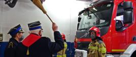 Widok z tyłu. Kapelan małopolskich strażaków w trakcie poświęcenia nowego pojazdu. Po lewej stronie aspirant PSP w tle dwóch strażaków JRG w ubraniach specjalnych stojących przed samochodem ratowniczo-gaśniczym. 