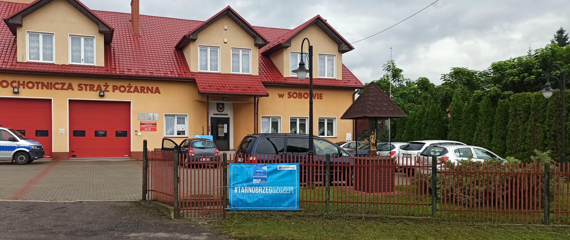 Budynek remizy OSP, przed którym stoją zaparkowane samochody osobowe. Na ogrodzeniu placu remizy znajduje się baner, który zachęca do zaszczepienia się przeciw COVID-19. 