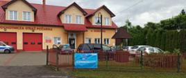 Budynek remizy OSP, przed którym stoją zaparkowane samochody osobowe. Na ogrodzeniu placu remizy znajduje się baner, który zachęca do zaszczepienia się przeciw COVID-19. 