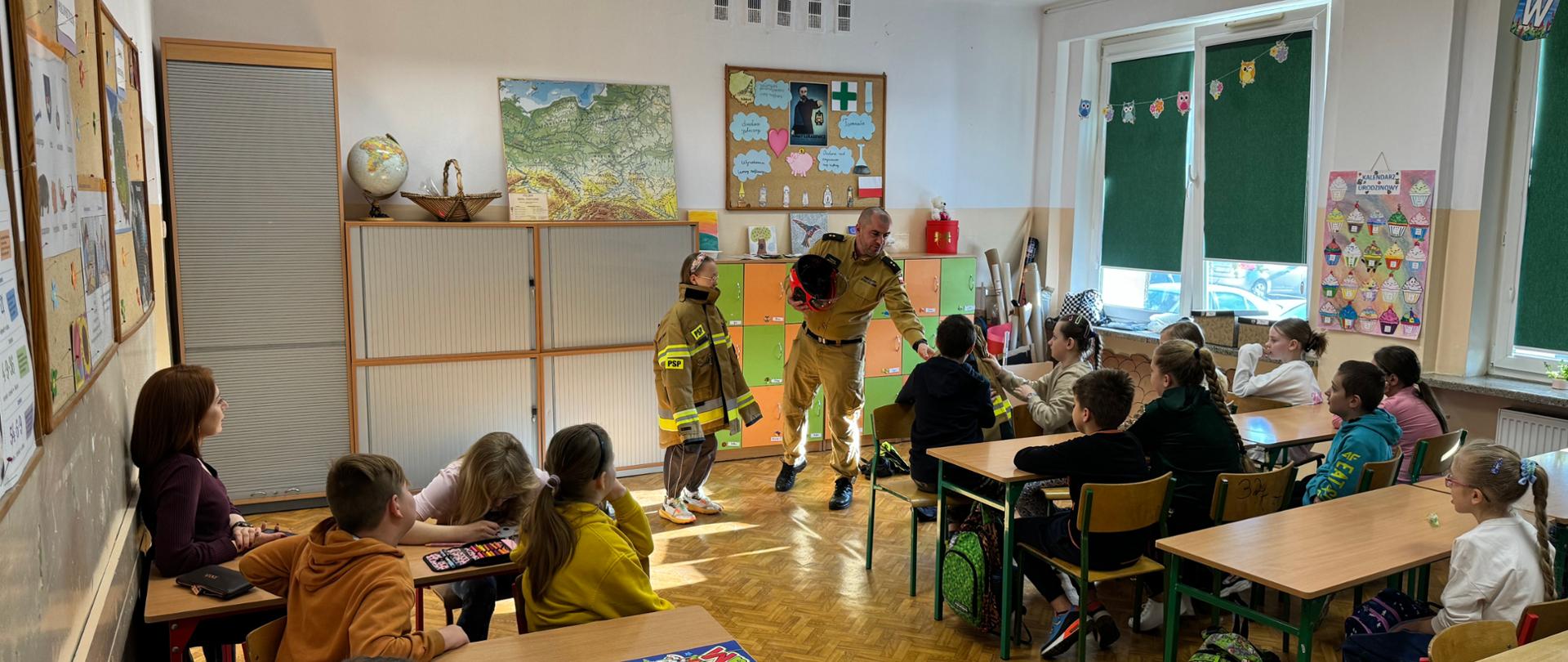 Strażak prowadzi zajęcia edukacyjne w Szkole Podstawowej nr 3 w Krośnie Odrzańskim. Prezentuje hełm i rozmawia z uczniami, z których jedno dziecko jest przebrane w strój strażacki. Pozostali uczniowie siedzą przy ławkach i uważnie słuchają.