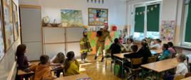 Strażak prowadzi zajęcia edukacyjne w Szkole Podstawowej nr 3 w Krośnie Odrzańskim. Prezentuje hełm i rozmawia z uczniami, z których jedno dziecko jest przebrane w strój strażacki. Pozostali uczniowie siedzą przy ławkach i uważnie słuchają.
