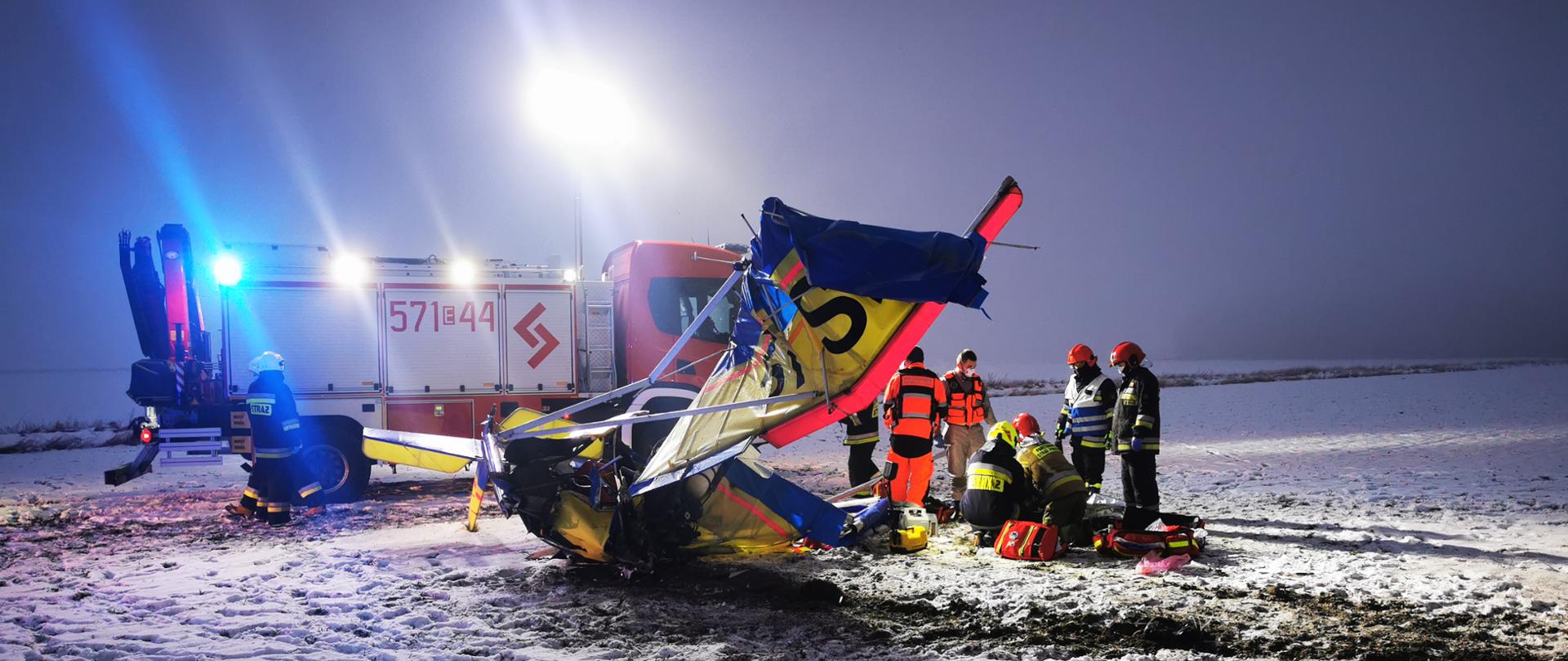 Zdjęcie przedstawia wrak samolotu ultralekkiego w m. Szadek, z prawej strony samolotu widoczni strażacy udzielający pomocy osobie poszkodowanej wraz z ratownikami medycznymi. W tle widać samochód strażacki oświetlający teren akcji. Na polu leży śnieg i porozrzucane są części samolotu. 