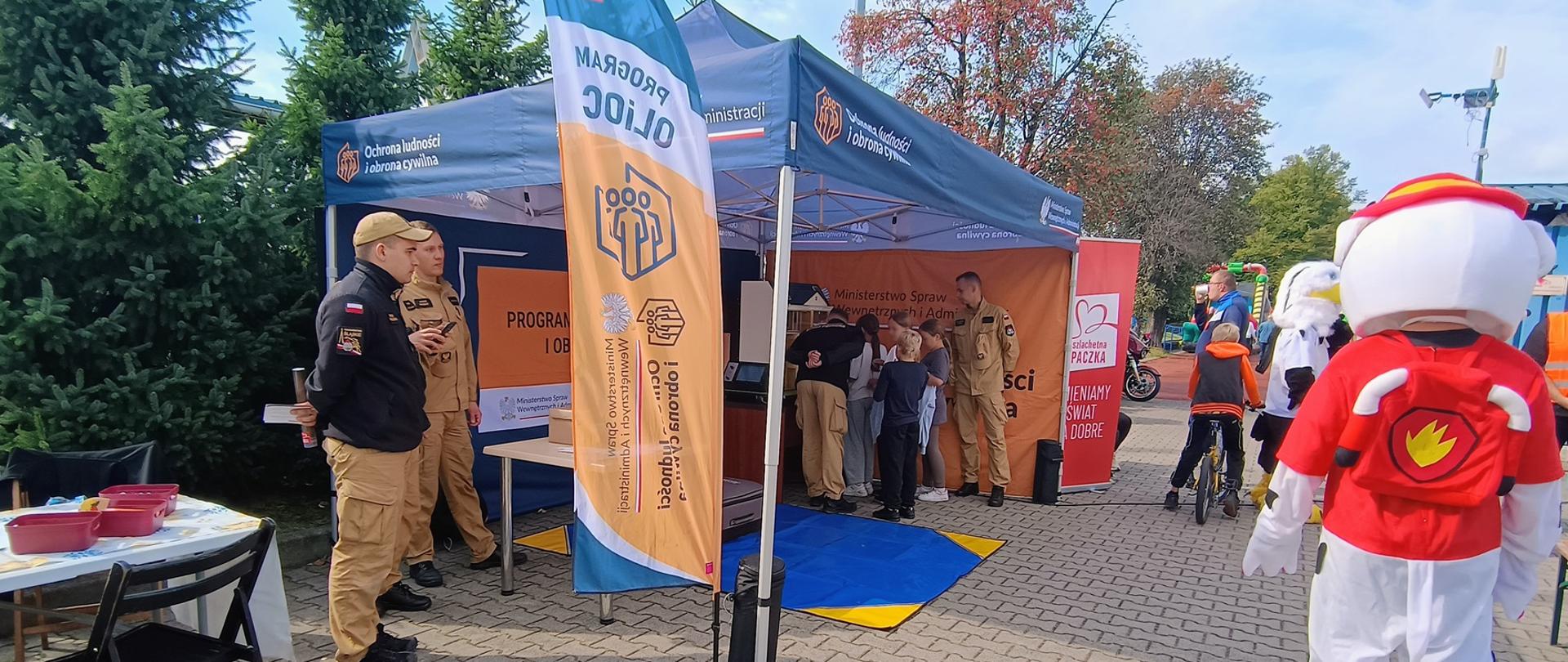 Zdjęcie przedstawia stoisko promocyjno-edukacyjne zorganizowane na świeżym powietrzu podczas festynu. Centralnym elementem jest niebieski namiot ze znakami i napisami odnoszącymi się do ochrony ludności i obrony cywilnej oraz Ministerstwa Spraw Wewnętrznych i Administracji. Obok stoi pionowy flaga-banner z napisem „PROGRAM OLIOC”. Przed namiotem oraz w jego wnętrzu znajdują się osoby w mundurach służb mundurowych oraz odwiedzający, w tym dzieci i dorośli rozmawiający ze stanowiskowymi instruktorami.
Na pierwszym planie po prawej stronie uwagę przyciąga osoba w dużym kostiumie maskotki – białego psa w czerwonym ubraniu strażackim z plecakiem z symbolem płomienia. W tle po prawej stronie widoczni są inni uczestnicy wydarzenia, m.in. dzieci na rowerach oraz maskotka orła. Z kolei po lewej stronie znajduje się stół nakryty obrusem z czerwonymi pojemnikami, przy którym ustawiono czarne krzesła. Całość wydarzenia odbywa się na wybrukowanym placu w otoczeniu zieleni i otwartej przestrzeni.
