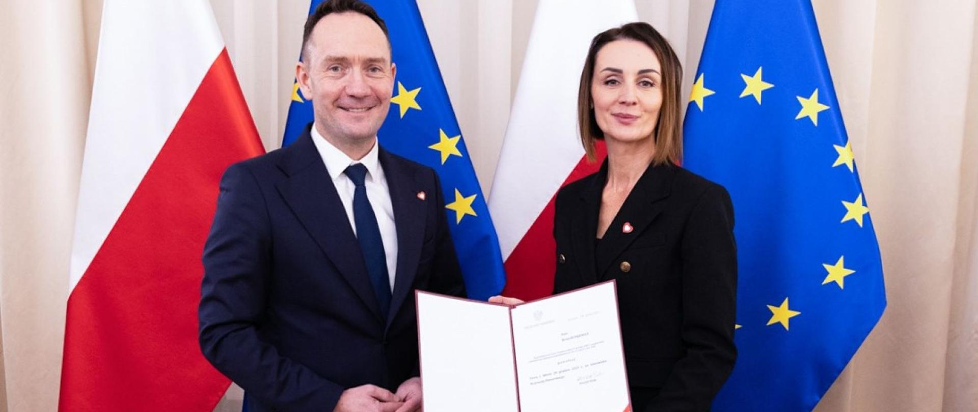Zdjęcie przedstawia dwie osoby, kobietę i mężczyznę. W tle widać flagę Polski i UE