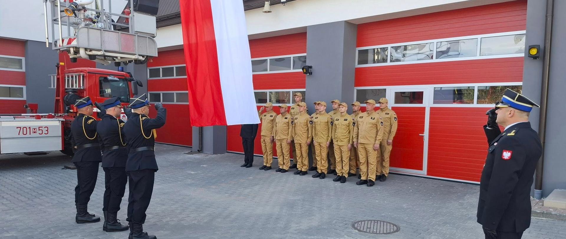 Zdjęcie przedstawia uroczystą ceremonię Państwowej Straży Pożarnej w Zwoleniu z okazji Dnia Flagi Rzeczypospolitej Polskiej. Grupa strażaków w mundurach stoi w szyku przed remizą strażacką z dużymi czerwonymi bramami garażowymi. Po lewej stronie widoczny jest wóz strażacki z drabiną. Na środku zdjęcia dumnie wisi duża biało-czerwona flaga Polski, ustawiona pionowo. Część strażaków salutuje fladze, co wskazuje na moment szacunku lub uroczystość.