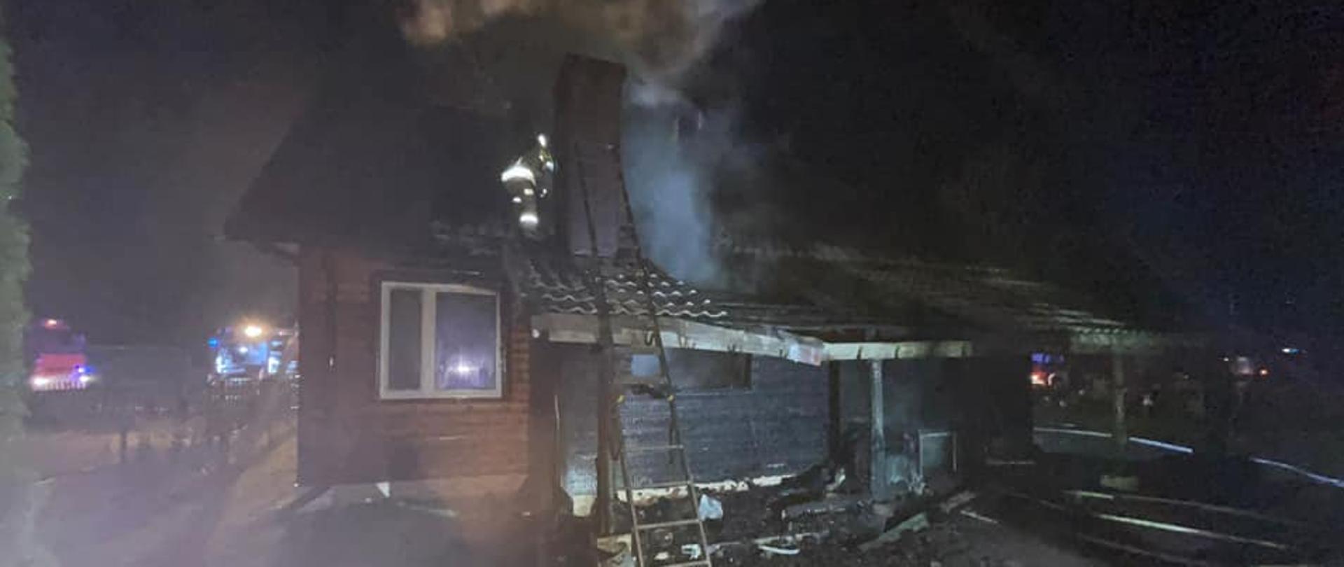 Budynek mieszkalny jednorodzinny po pożarze widziany od strony podwórka. Uszkodzona część dachu. Nadpalone ściany budynku. Do budynku przystawiona drabina strażacka. W tle samochody ratowniczo-gaśnicze.