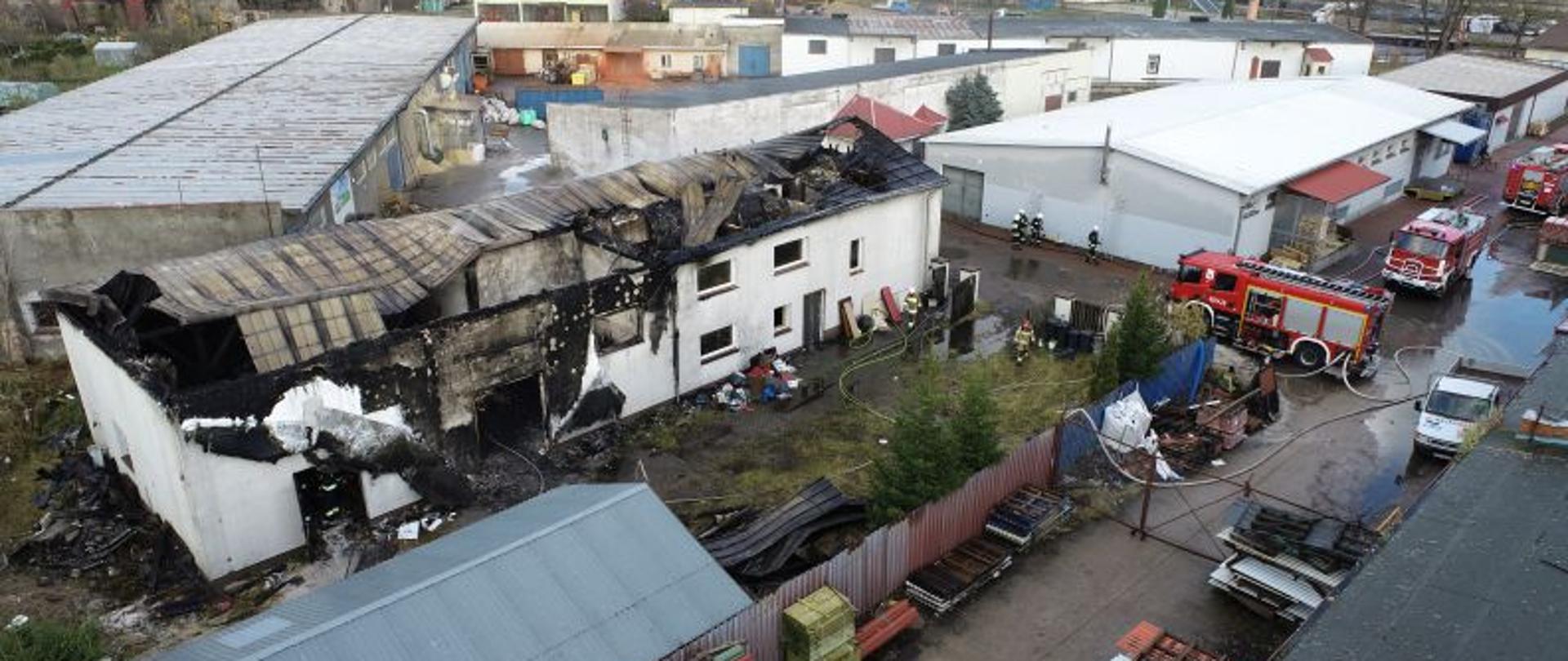 Zdjęcie wykonane z drona. W centralnej części uszkodzony, biały budynek. Budynek po pożarze. Mam spalony i zapadnięty dach. Do około strażacy, wozy bojowe straży pożarnej i porozbijane węże