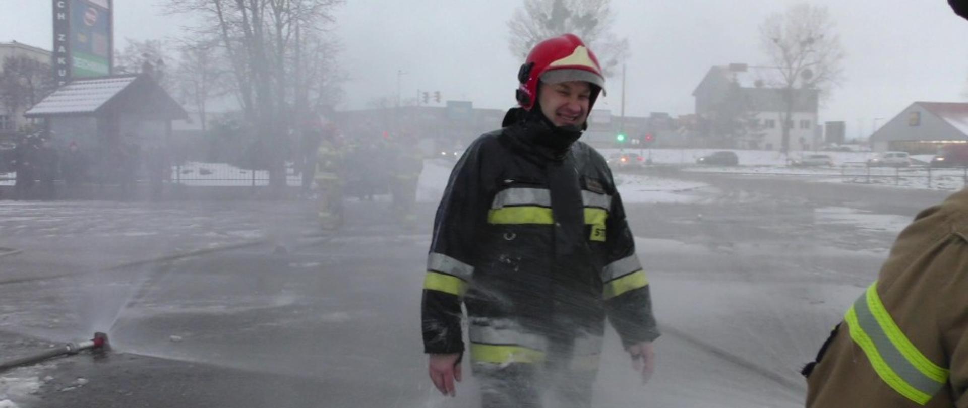 Na zdjęciu strażak ubrany w ubranie specjalne oraz hełm, na którego leje się woda z kurtyn wodnych.