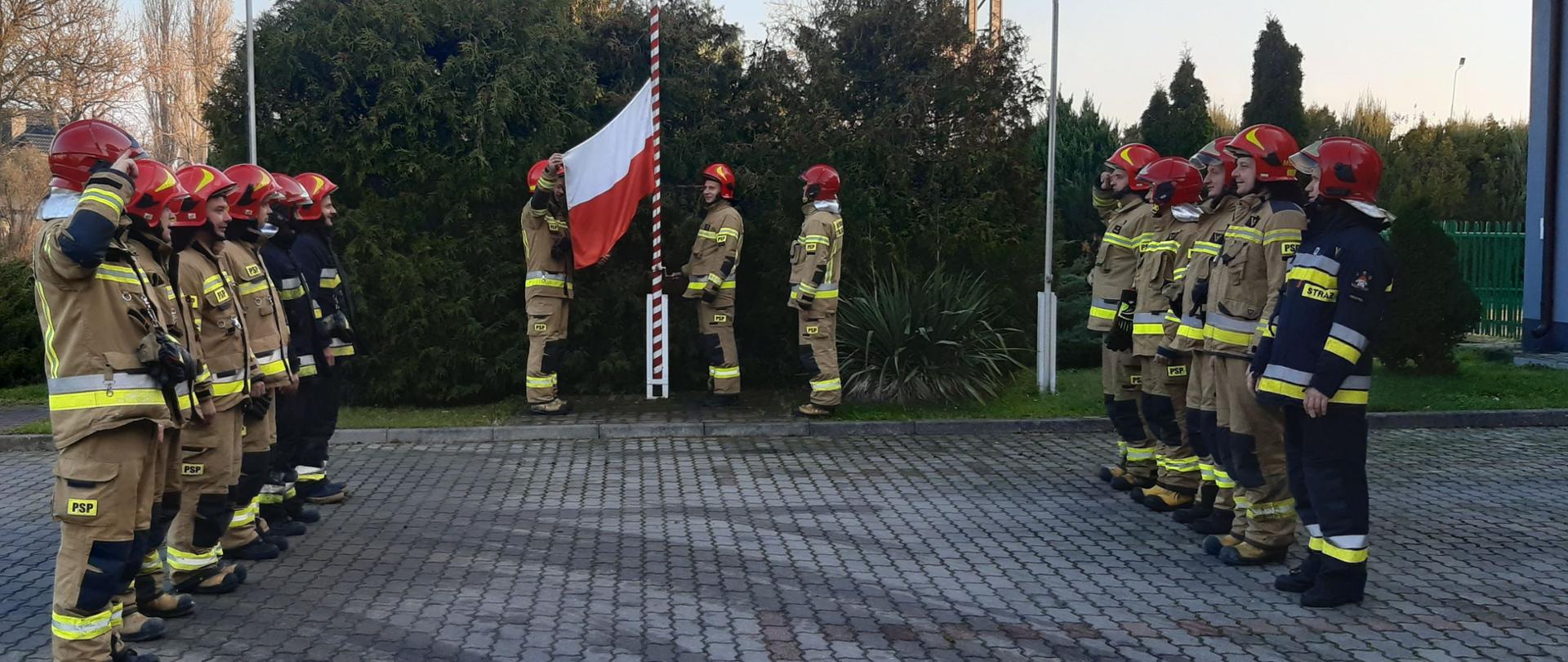 Na zdjęciu widać strażaków w żółtych i granatowych strojach ora czerwonych hełmach. W dwóch rzędach naprzeciwko siebie stoi jedenastu strażaków (sześciu w lewej stronie i pięciu po prawej). Następnych trzech strażaków w żółtych strojach i czerwonych hełmach zawiesza flagę, która będzie wciągana na maszt. W tle widać wspinalnie oraz krzaki. Z prawej strony widać kawałek budynku Komendy Powiatowej Państwowej Straży Pożarnej w Łęczycy. Jest widno.