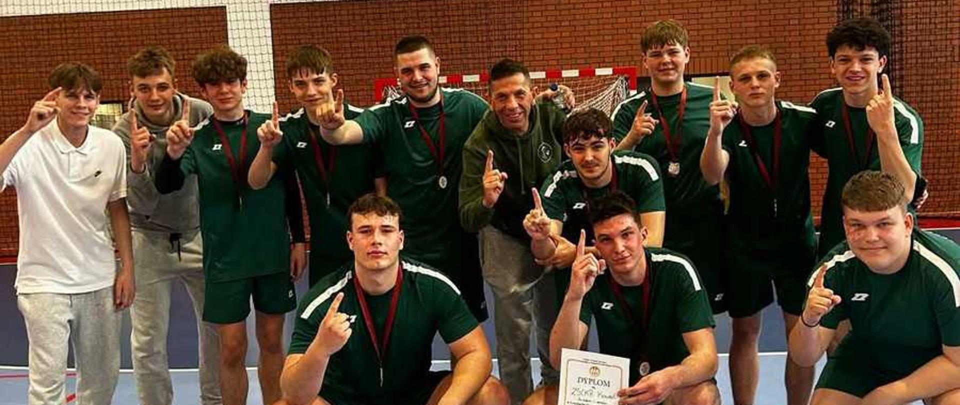 Zdjęcie przedsatawia drużynę koszykarską wraz z trenerem. Fotografia zrobiona jest na nowowczesnej hali sportowej. Młodziez ubrana zw stroje barwy zielonej. Trener ma założoną bluze z napisam Zespół Szkół Centrum Kształcenia Rolniczego w Kowalu. Przed drużyną, na podłodze stoi puchar oraz dyplom mówiące o zajęciu I miejsca przez powyższą drużynę.