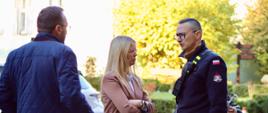 Trzy osoby rozmawiające na placu. Po prawej stronie stoi strażak w czarnej bluzie mundurowej z radiostacją i okularach, trzymający kartkę papieru. Naprzeciwko niego stoi kobieta w różowo-beżowej marynarce z założonymi rękami, a po lewej mężczyzna w pikowanej niebieskiej kurtce odwrócony tyłem do obiektywu.
