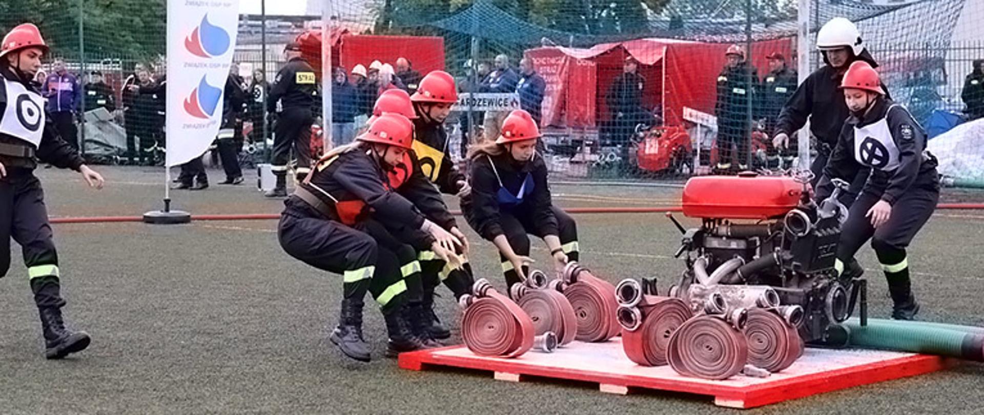 na podeście znajduje się motopompa sześć węży i prądownica cztery kobiety podbiegają i chwytają czerwone węże mężczyzna z kobietą podbiegają do motopompy w tle znajduje się liczna widownia