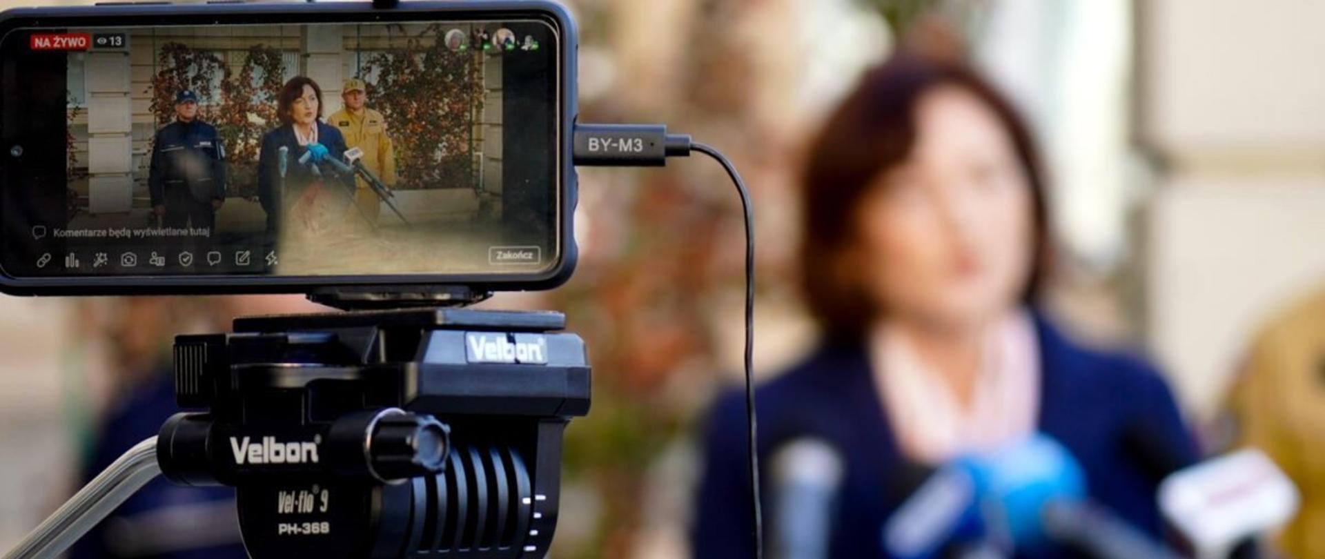 Zdjęcie zrobione na zewnątrz, w trakcie briefingu prasowego. Na ekranie smartfona, który służy do prowadzenia transmisji online briefingu prasowego widać mówiącą do mikrofonów wojewodę podkarpackiego. Z tyłu stoją oficerowie Policji i Państwowej Straży Pożarnej.