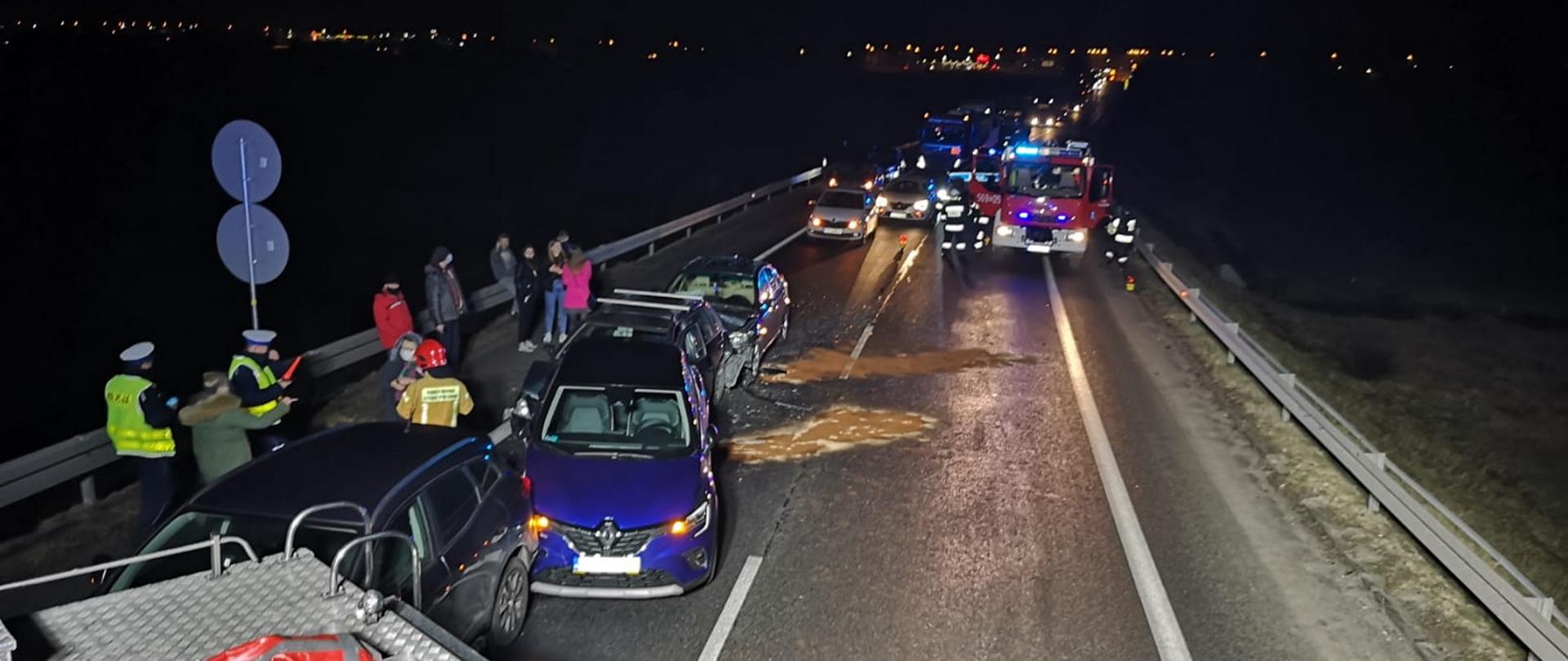 Zdjęcie ze zdarzenia komunikacyjnego. Na drodze stoją rozbite samochody a tuż obok nich uczestnicy zdarzenia oraz Funkcjonariusze Państwowej Straży Pożarnej oraz Policji. Na jezdni jest zneutralizowany wyciek płynów eksploatacyjnych. W tle zdjęcia widać tworzący się korek. 