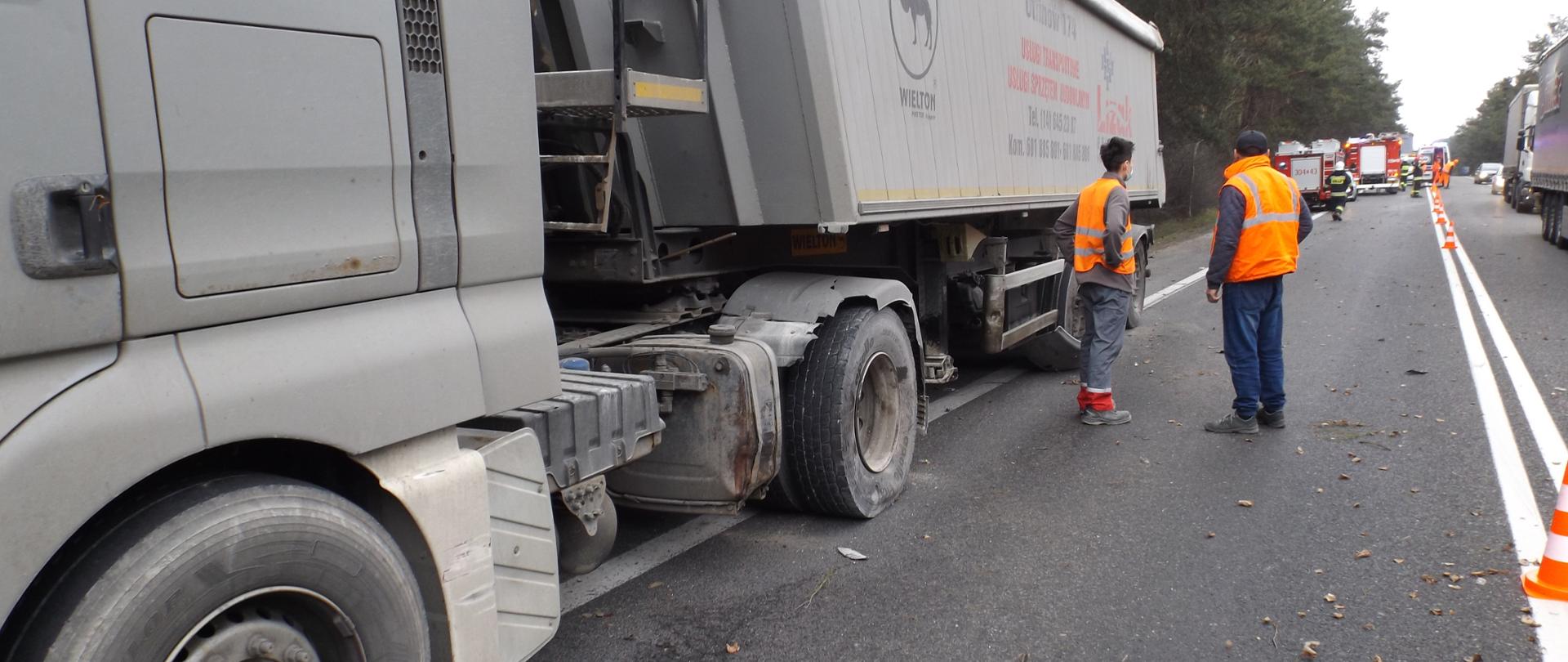 Na zdjęciu znajduje się samochód ciężarowy z lekkimi uszkodzeniami koła oraz nadkola. W tle widać samochody strażackie, oznakowaną pachołkami jezdnię, oraz kierowcę samochodu ciężarowego w odblaskowej kamizelce 