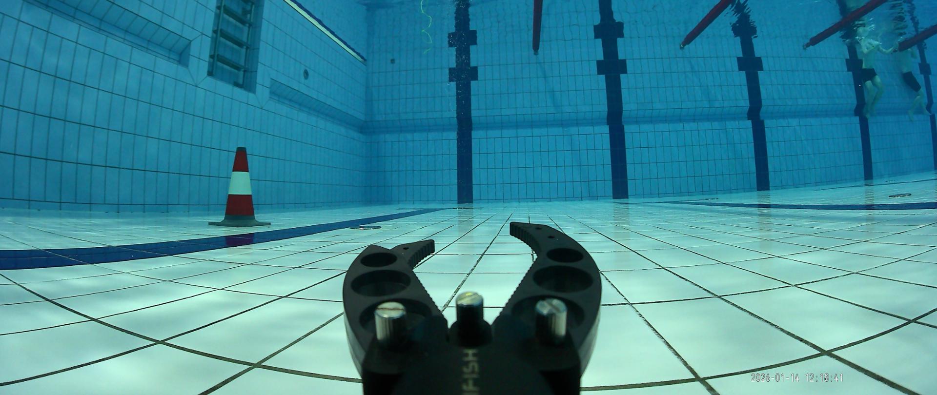 Widok dna basenu z perspektywy podwodnej; na pierwszym planie znajduje się chwytak robota podwodnego, w tle pachołek treningowy i linie torów pływackich