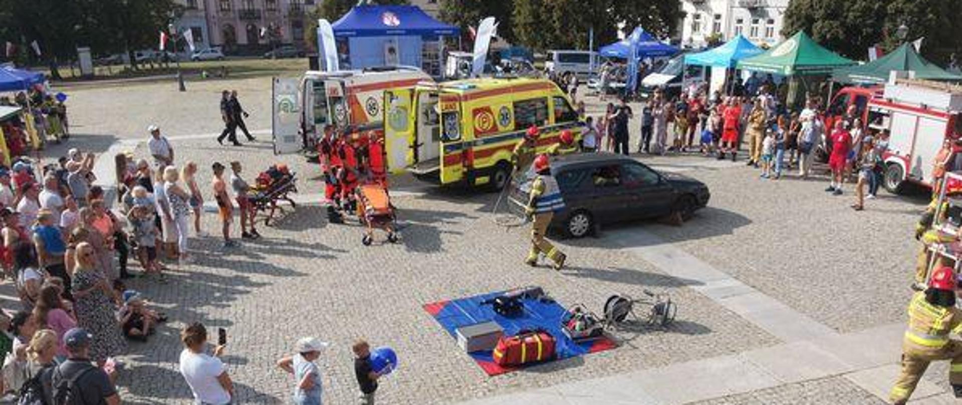 Zdjęcie wykonane z drona podczas pikniku na rynku w Radomiu. Na zdjęciu widać pokaz symulowanego wypadku drogowego wykonywany przez strażaków. Dookoła miejsca pokazu stoją osoby postronne przybyłe na piknik. 