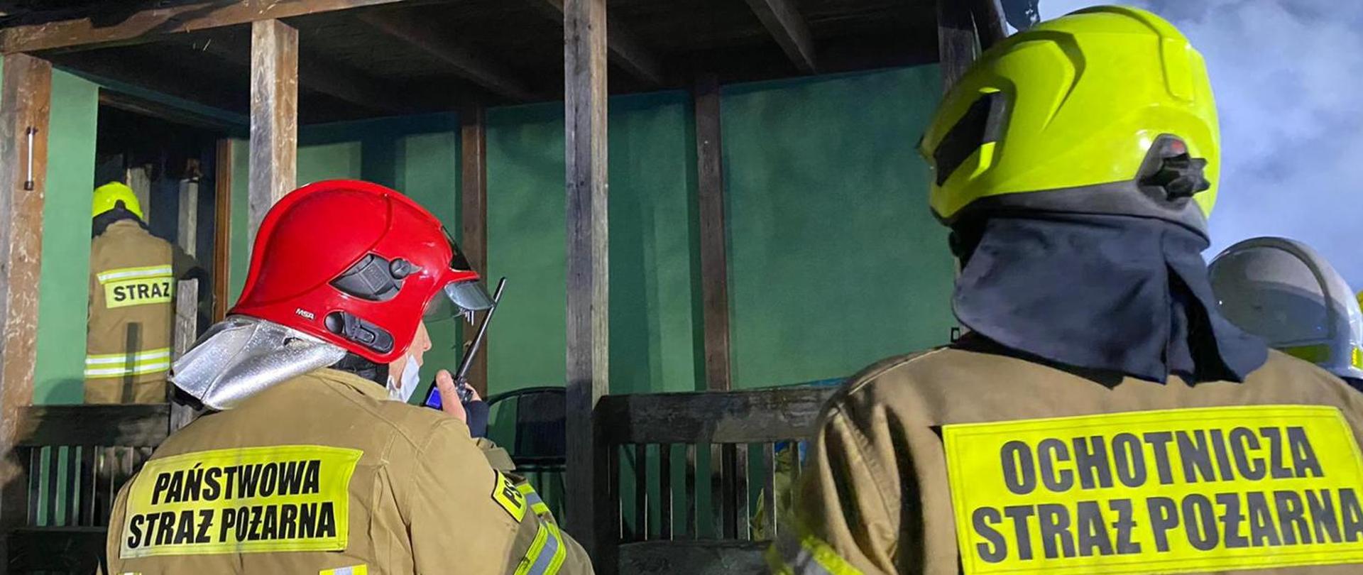 Zdjęcie przedstawia Funkcjonariusza Państwowej Straży Pożarnej oraz Ochotniczej Straży Pożarnej odwróconych tyłem w umundurowaniu bojowym. 