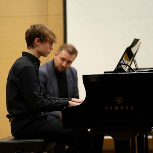 zdjęcie przedstawia ucznia grającego na jednym z dwóch fortepianów na scenie sali koncertowej, w tle prowadzący warsztaty przyglądający się w grze ucznia