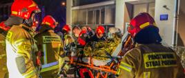 Na tym zdjęciu widać kilku strażaków oraz ratowników medycznych, transportujących poszkodowaną osobę z poparzeniami do karetki. Osoba przewożona jest na noszach, owinięta folią NRC. Poszkodowanemu udzielana jest tlenoterapia bierna. Część strażaków ubrana jest w AODO. Cała akcja ma miejsce w godzinach nocnych.