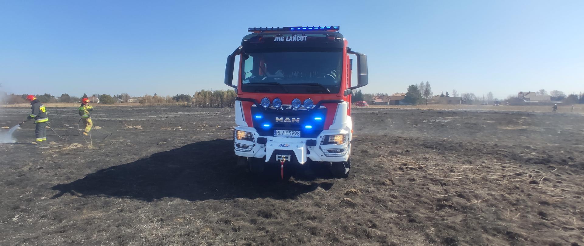 Zdjęcie przedstawia pozostałości po pożarze łąki na którym pracują strażacy dogaszając powstały pożar nieużytków. Na pierwszym planie średni samochód ratowniczo-gaśniczy MAN będący na wyposażeniu Komendy Powiatowej Państwowej Straży Pożarnej w Łańcucie