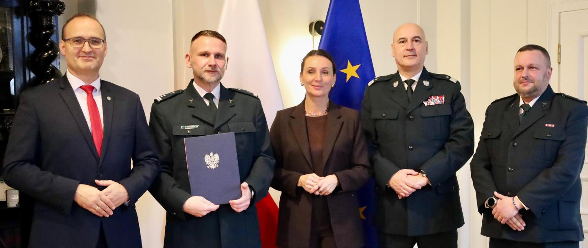 Zdjęcie przedstawia pięć osób - czterech mężczyzn i jedną kobietę - stojących razem w pomieszczeniu. W tle widoczne są flagi Polski i Unii Europejskiej. Mężczyzna po lewej stronie trzyma złożony dokument w niebieskiej okładce.