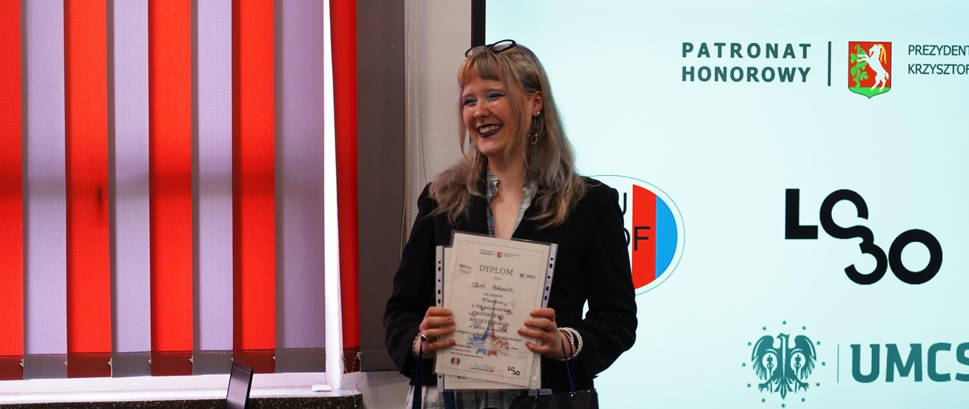 Rozpromieniona dziewczyna, Oliwia Petkowicz trzyma dyplom i torbę z logo UMCS, stojąc obok ekranu prezentującego logotypy organizatorów. Zdjęcie dokumentuje sukces uczestniczki w konkursie recytatorskim.