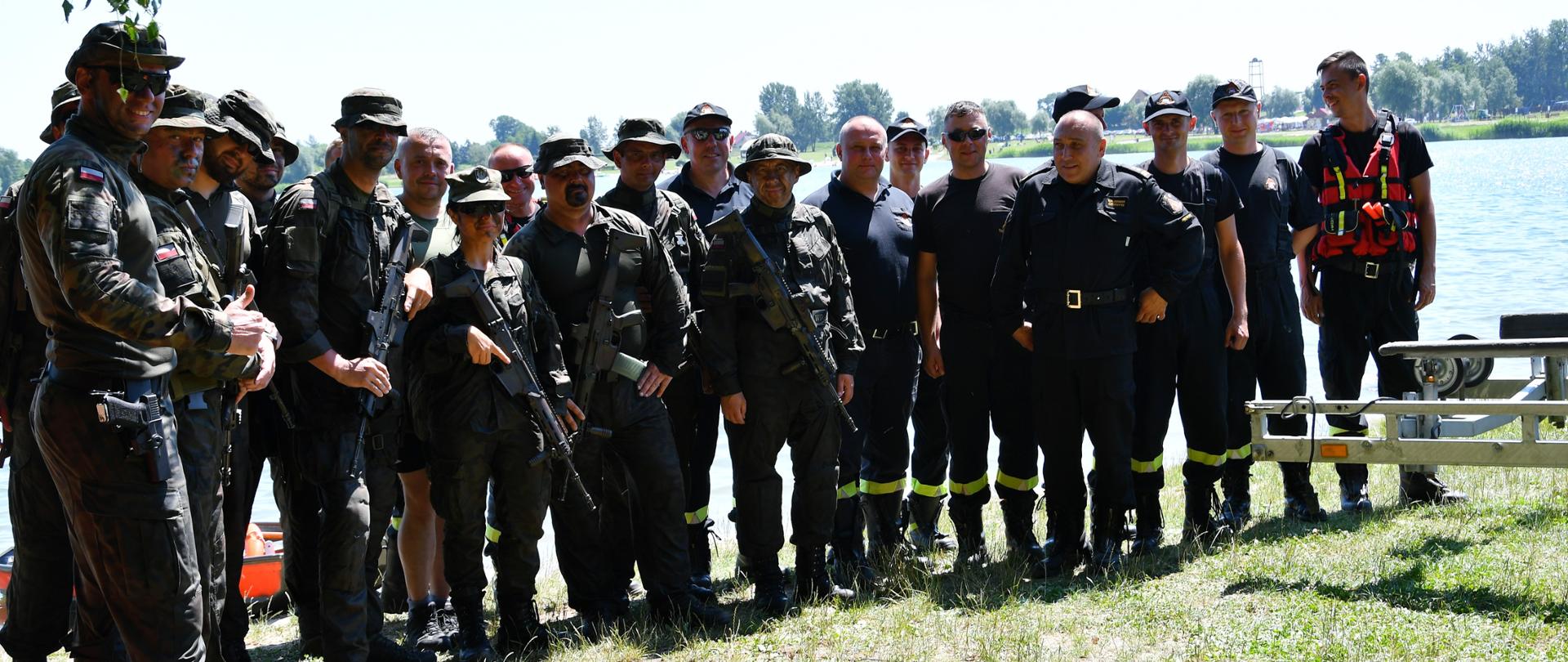 Zdjęcie grupowe strażaków oraz żołnierzy biorących udział we wspólnych ćwiczeniach. Wszystkie osoby ubrane są w umundurowanie koszarowe. W tle widoczny jest zbiornik wodny.