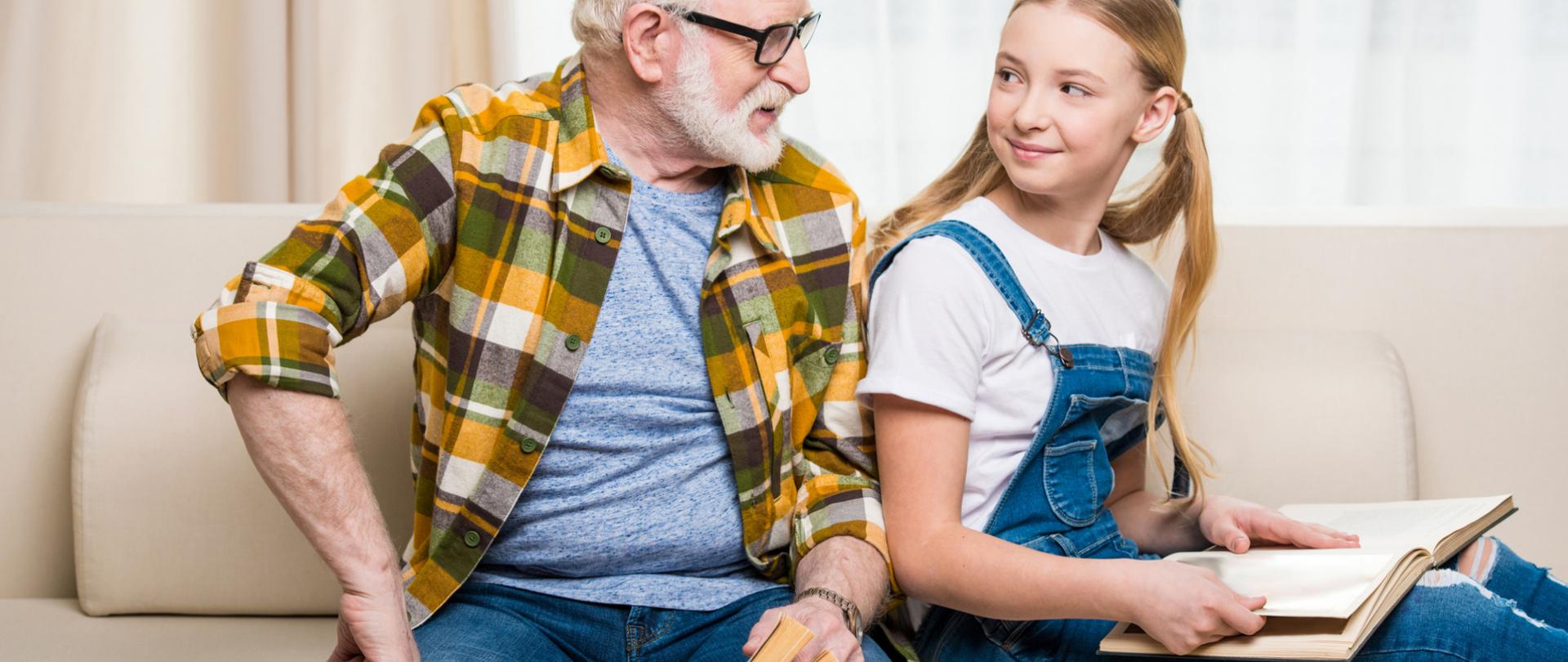 Na zdjęciu jest starszy mężczyzna wraz z młodą dziewczyną. Siedzą na kanapie trzymając w dłoniach książki.