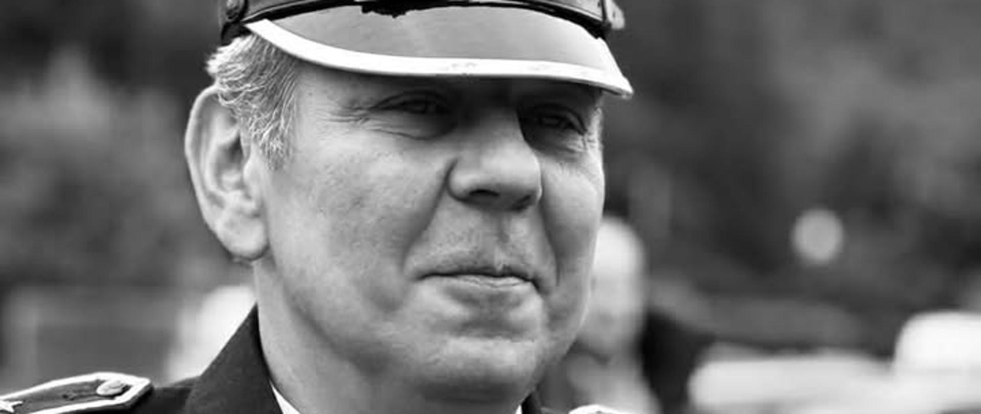 Czarno-białe zdjęcie portretowe mężczyzny w średnim wieku, ubranego w galowy mundur strażacki. Mężczyzna patrzy lekko w bok z delikatnym, życzliwym uśmiechem. Na głowie ma rogatywkę z orłem pośrodku oraz trzema gwiazdkami umieszczonymi nad daszkiem. Ubrany jest w ciemną marynarkę mundurową z pagonami, białą koszulę i ciemny krawat. Na kołnierzu widoczne są emblematy w kształcie stylizowanych gwiazd (ogników), a na piersi znajduje się plakietka z nazwiskiem „A. PAWLAK”. Zdjęcie przedstawia zmarłego aspiranta sztabowego w stanie spoczynku Artura Pawlaka, który był oddanym i godnym naśladowania strażakiem.