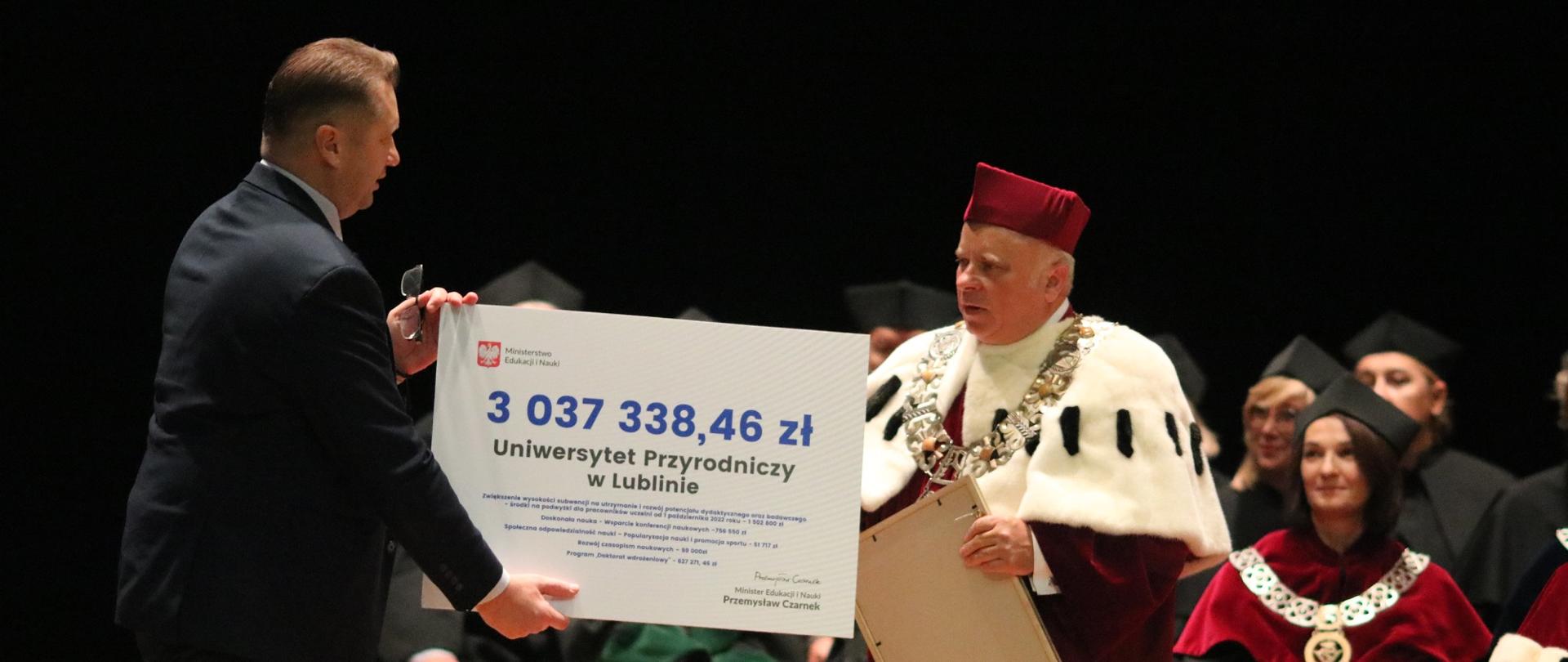 Minister wręcza mężczyźnie w gronostajowym płaszczu wielki symboliczny czek z napisem 3 037 338,46 zł.