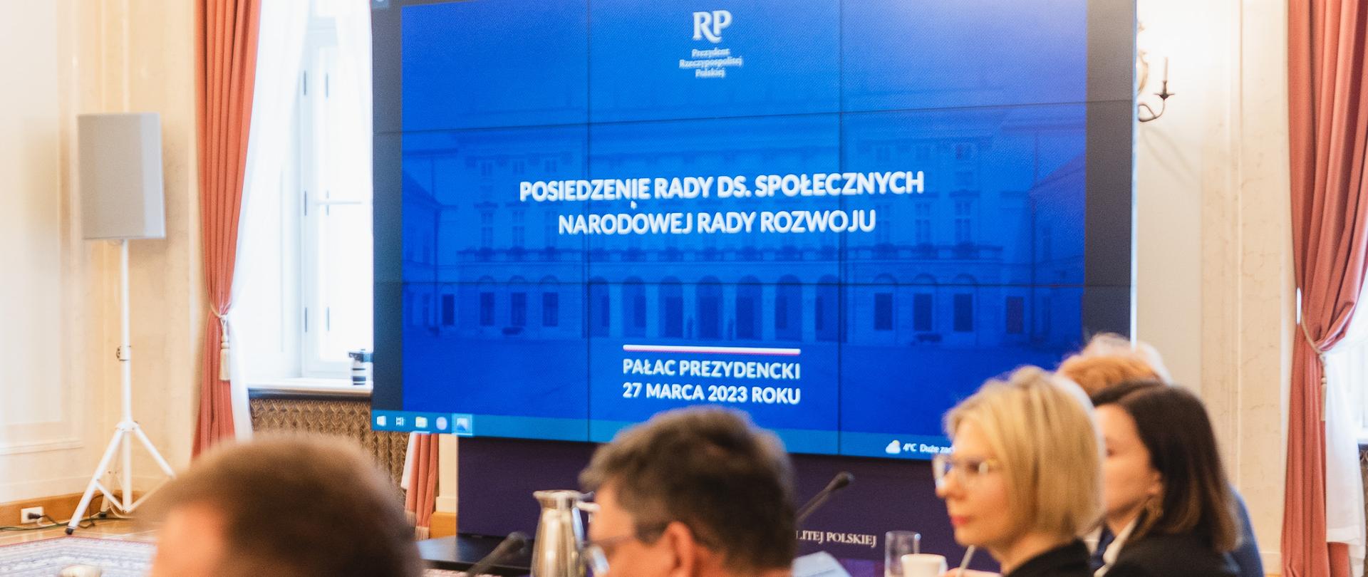 Na pierwszy planie osoby siedzące przy stole. W tle ekran z prezentacją