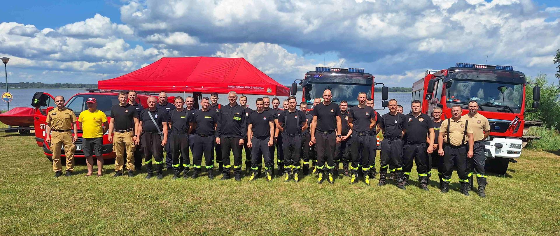 Zdjęcie grupowe uczestników ćwiczeń. Osoby stoją w dwuszeregu, w tle pojazdy pożarnicze i zbiornik wodny.