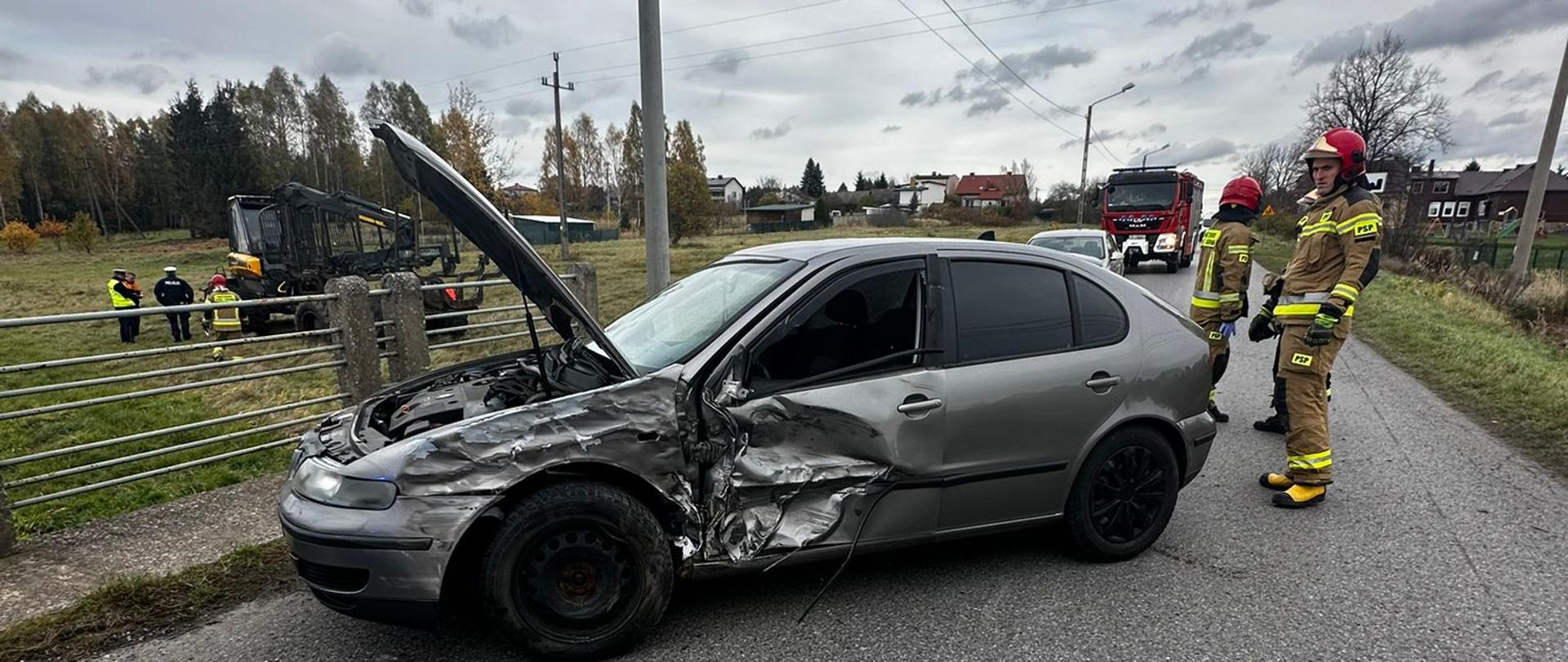 Samochód osobowy po kolizji z pojazdem przeznaczonym do transportu drewna (forwarderem). Obok samochodu stoją strażacy. W tle widać pojazd do transportu drewna i stojących przy nim strażaków i policjanta.