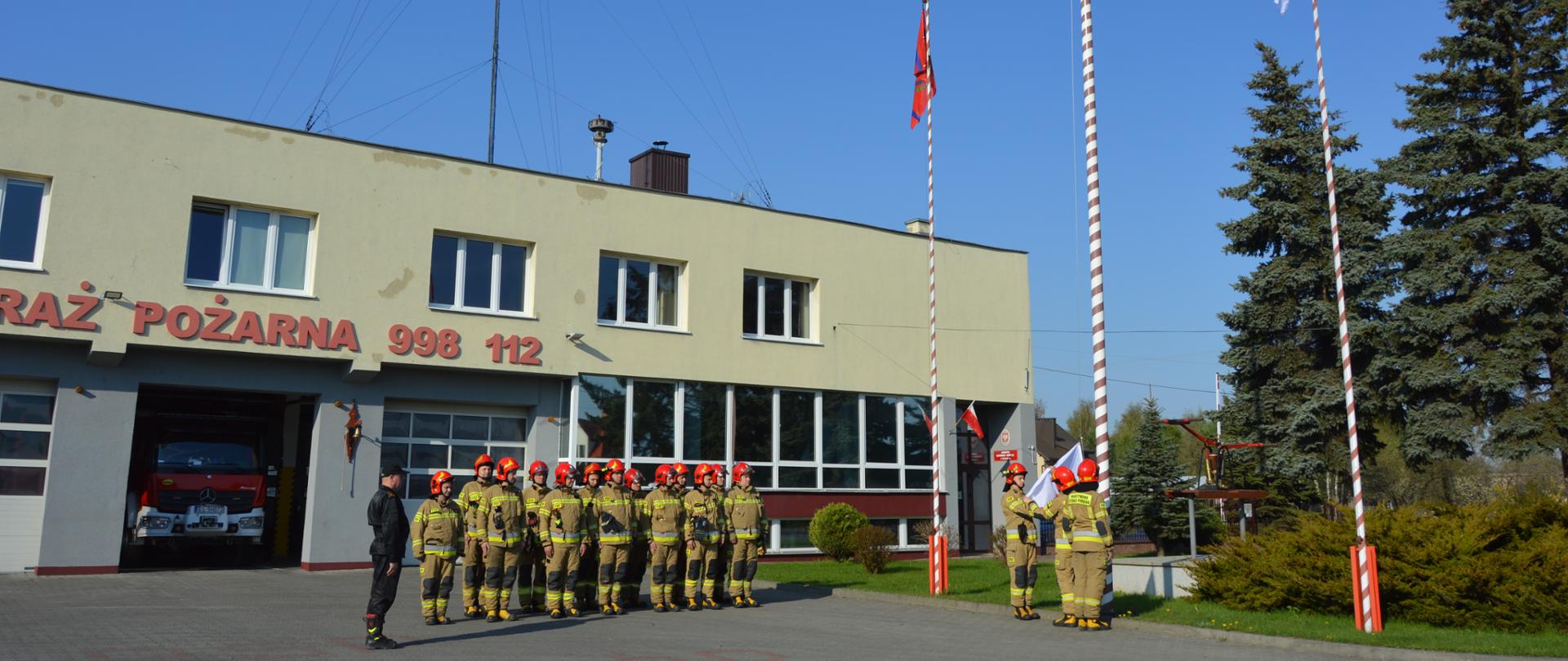 Zdjęcie przedstawia strażaków Komendy Powiatowej PSP powiatu łódzkiego wschodniego podczas uroczystego wciągnięcia flagi państwowej na maszt. Na zdjęciu widoczni strażacy ustawieni w dwuszeregu w jaskrawych ubraniach bojowych według nowego wzoru, w czerwonych hełmach. Z lewej strony salutuje dowódca. Z prawej strony przy maszcie flagowym salutuje trzech strażaków pocztu flagowego. Zdjęcie wykonane na tle strażnicy Jednostki Ratowniczo-Gaśniczej w Koluszkach. 