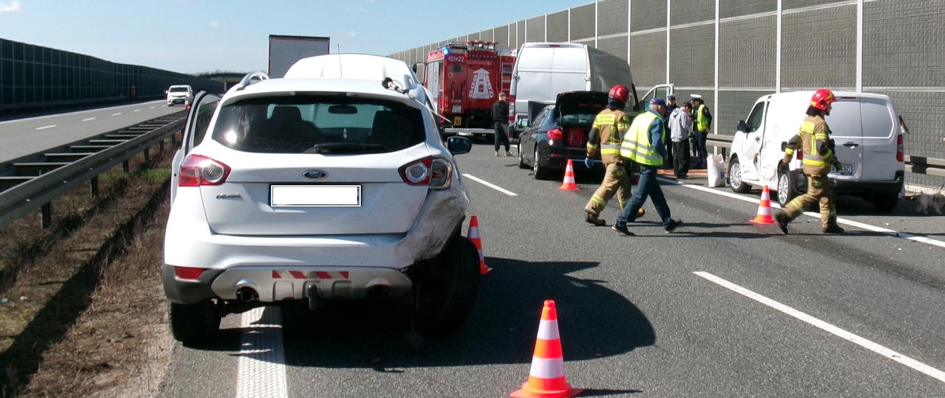 Zdjęcie z miejsca wypadku. Widoczne z cztery tyłu pojazdy biorące udział w zdarzeniu. W tle widać pojazd straży pożarnej. Ponadto widać strażaków zabezpieczających miejsce zdarzenia oraz inne osoby stojące w pobliżu uszkodzonych pojazdów. 