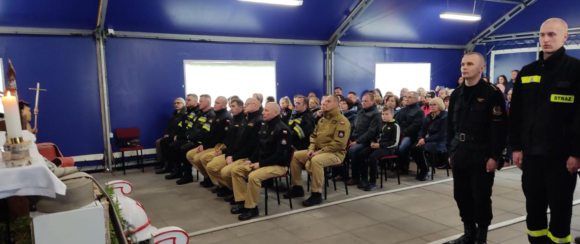 Nabożeństwo na terenie Przeciwpożarowej Bazy Leśnej Szkoły Głównej Służby Pożarniczej (PBL SGSP) w Zamczysku Nowym. Zdjęcie przedstawia namiot z ołtarzem i rzędami krzesełek na których siedzą studenci i kadra SGSP. 
