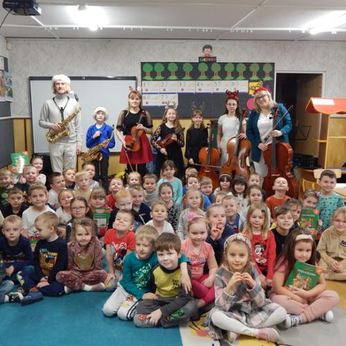 Na zdjęciu sala przedszkolna w Przedszkolu Miejskim nr 4 w Grajewie. Na pierwszym planie dzieci-widzowie siedzący po turecku, za nimi wykonawcy koncertu "Świąteczne smyki i stroiki czyli zabawa z saksofonem, skrzypcami i wiolonczelą". Wykonawcy maja ze sobą instrumenty, wiolonczele, skrzypce, saksofon. Na głowach mają czapki Mikołaja i inne ozdoby świąteczne. W tle po lewej stronie meble i zabawki przedszkolne, na środku monitor interaktywny, oraz tablica, na której zawieszone są wycięte z kolorowego papieru drzewa z namalowanymi jabłkami, pod nimi powieszone obrazki z cyframi, po prawej stronie drewniany stragan-zabawka, stoły i krzesła.