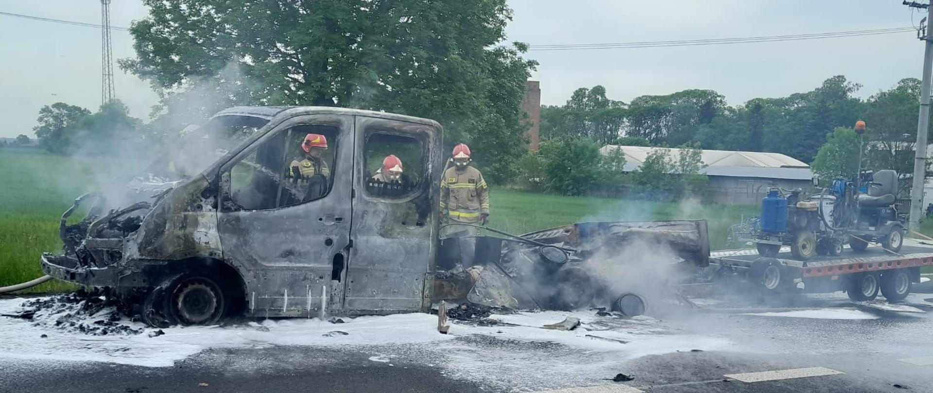 Na zdjęciu widać wrak spalonego samochodu dostawczego z przyczepą, który stoi na jezdni. Na przyczepie stoi jakaś maszyna na kółkach. Za samochodem widać trzech strażaków w żółtych strojach i czerwonych hełmach. Na jezdni jest piana gaśnicza. W tle widać budynki, słupy energetyczne, drzewa oraz pole. Jest widno.