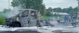 Na zdjęciu widać wrak spalonego samochodu dostawczego z przyczepą, który stoi na jezdni. Na przyczepie stoi jakaś maszyna na kółkach. Za samochodem widać trzech strażaków w żółtych strojach i czerwonych hełmach. Na jezdni jest piana gaśnicza. W tle widać budynki, słupy energetyczne, drzewa oraz pole. Jest widno.